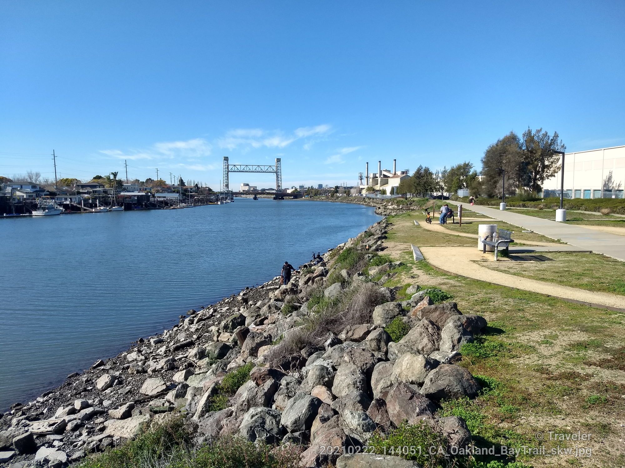 A river with a bridge, rocky bank, and people on a sunny path. - Oyster Bay Regional Shoreline – Oyster Bay Regional Shoreline loop from San Leandro