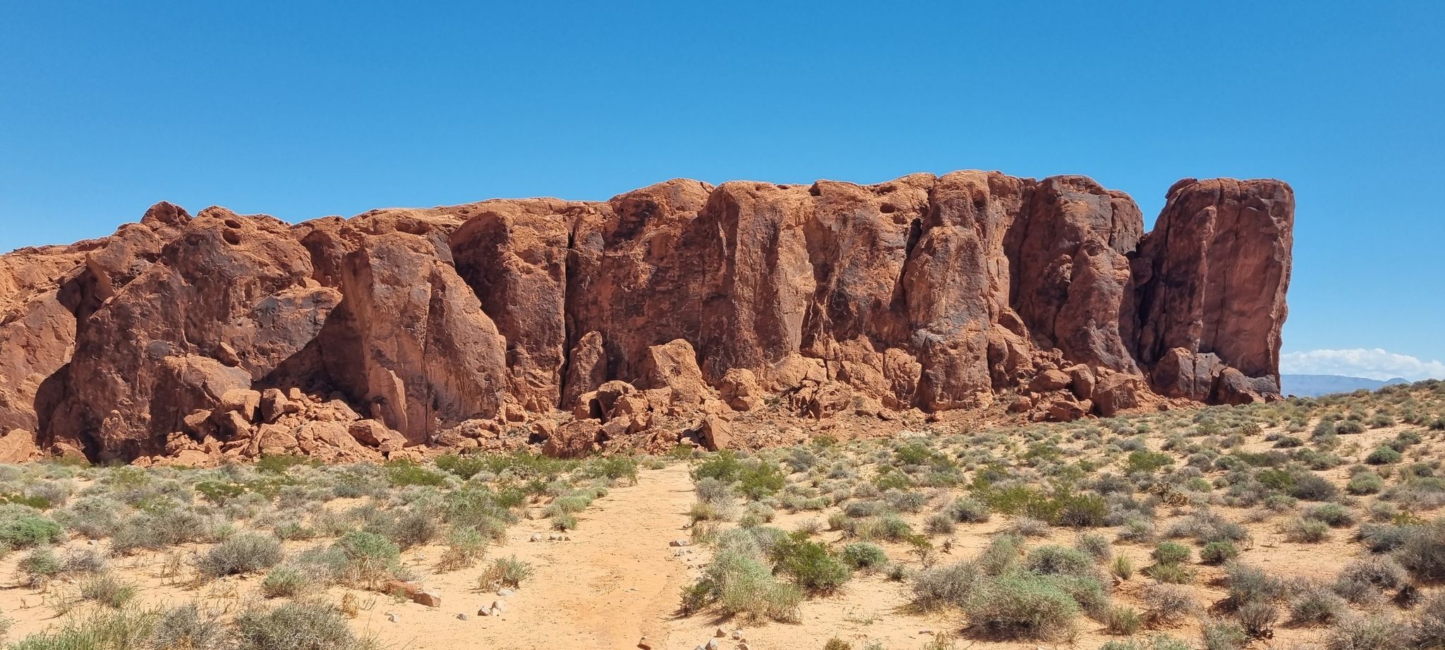 Long red rock wall with a desert path and shrubs, clear blue sky. - Fire Wave Trail