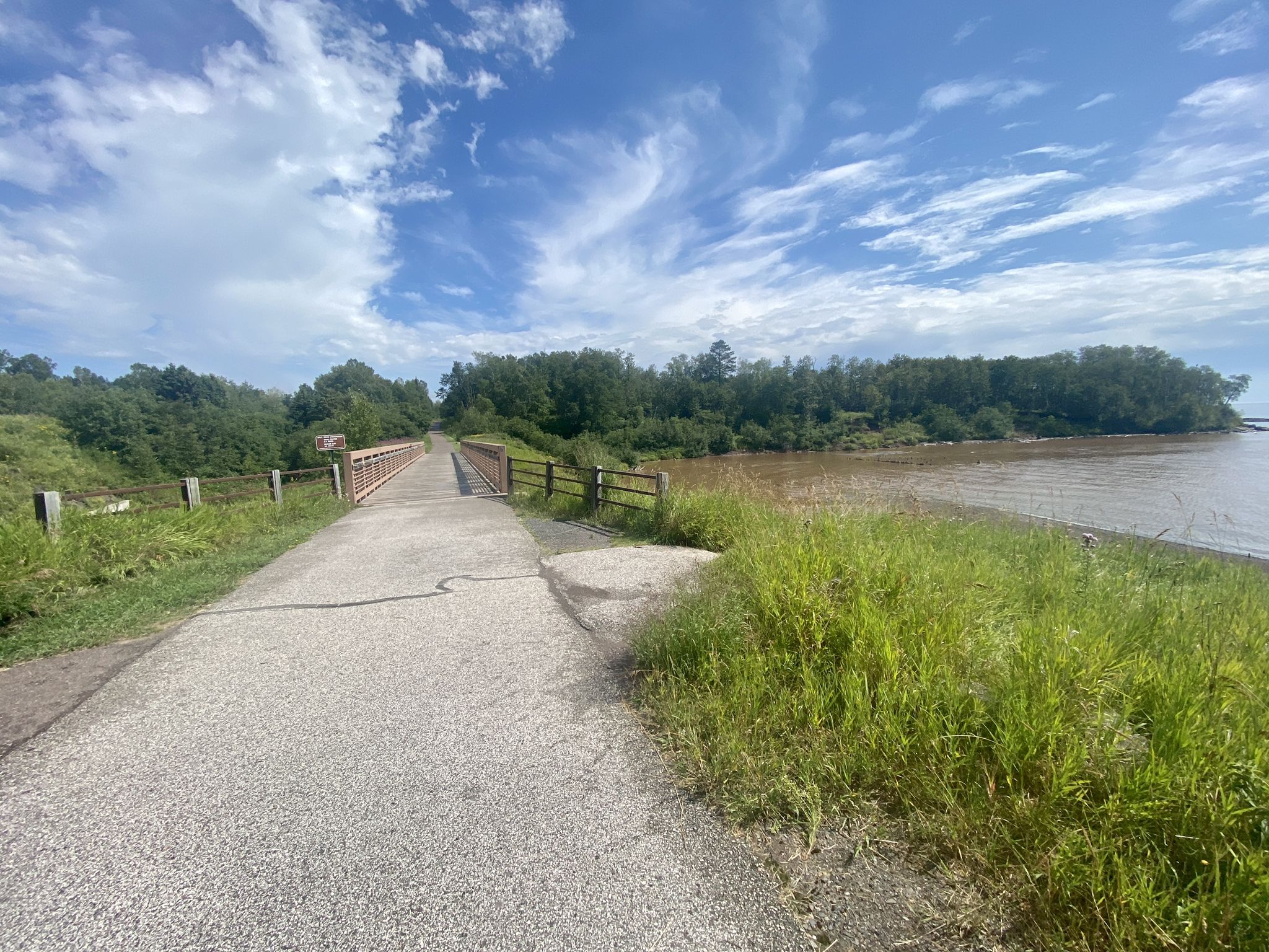 A paved path leads to a metal bridge over calm water, surrounded by green trees. - Gitchi-Gami Trail loop run
