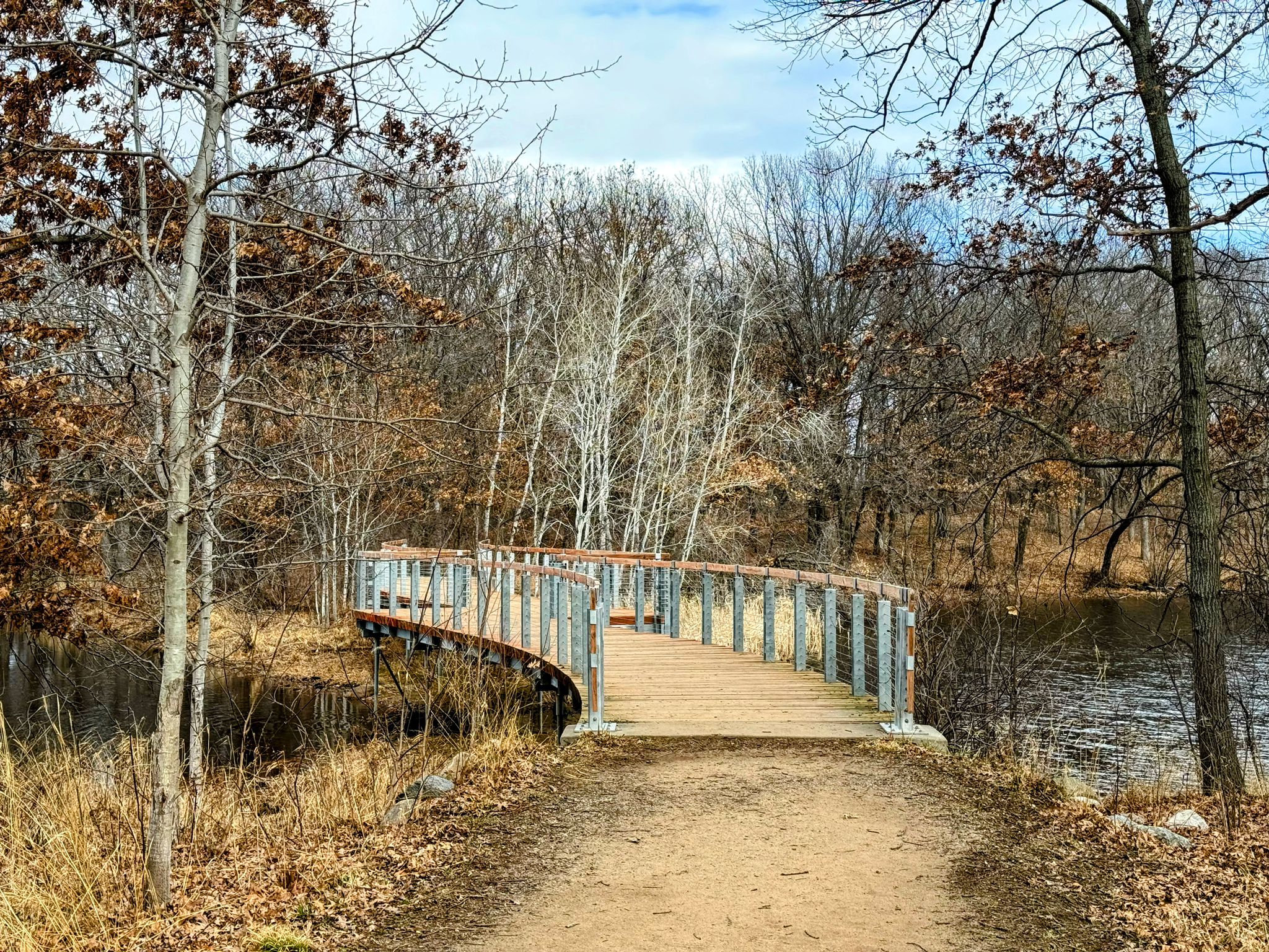 Curved wooden bridge over water, surrounded by bare trees under a cloudy sky. - Voyageur Trek Loop