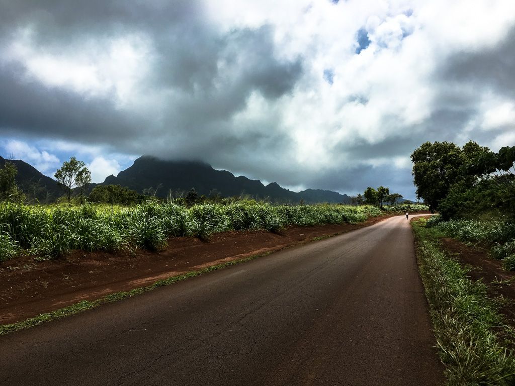An empty road stretches towards distant mountains under a cloudy sky. - Menehune Fishpond loop from Lihue