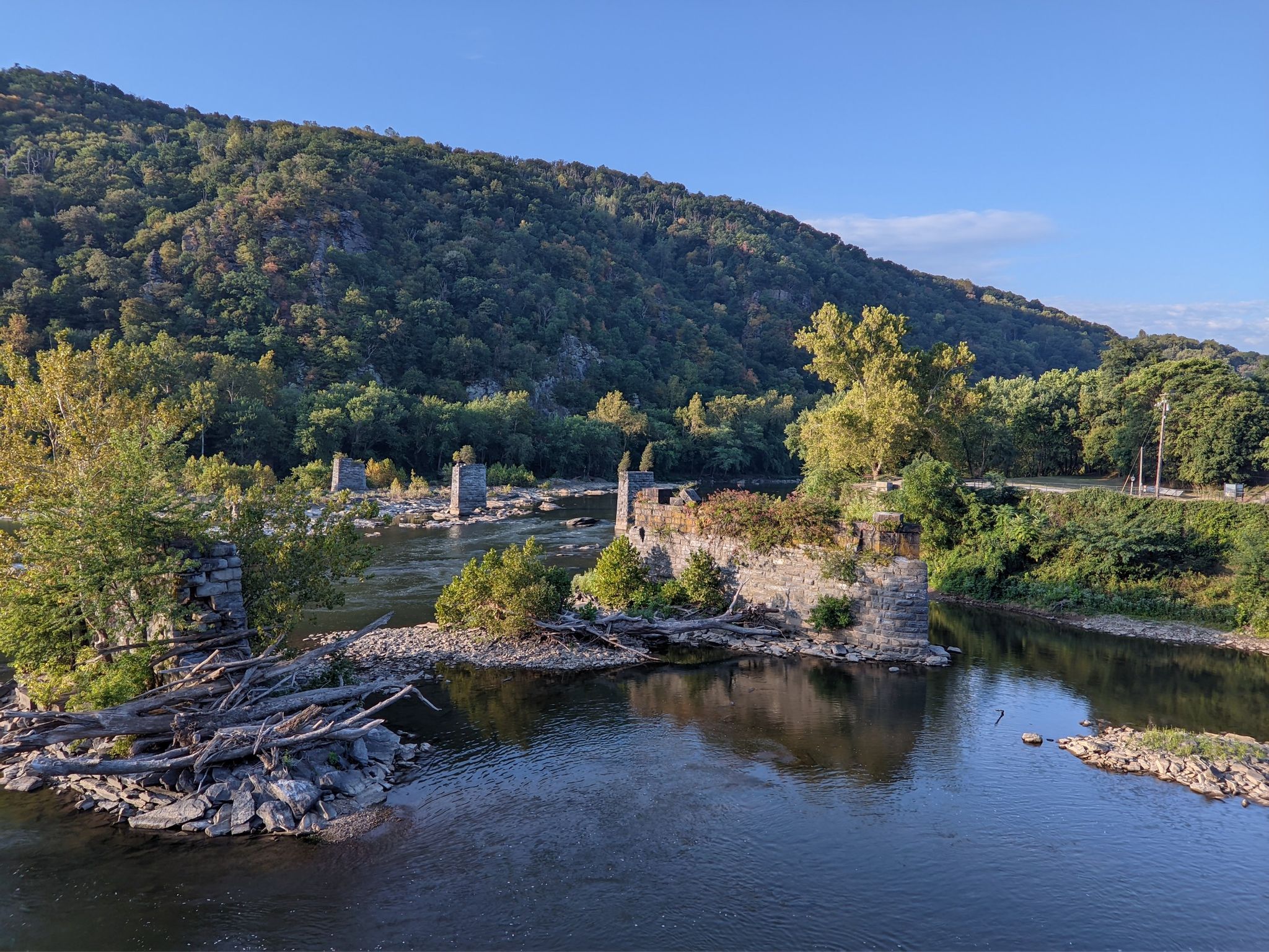 River with stone ruins, green trees, and forested mountains under clear sky. - Chesapeake and Ohio Canal Towpath