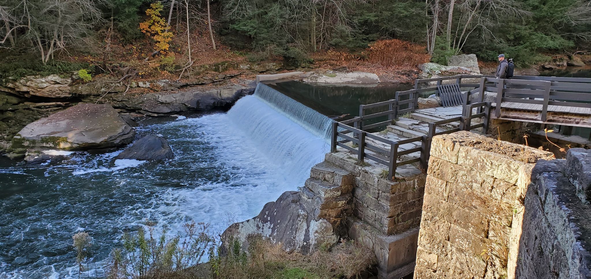 A person stands on a wooden bridge overlooking a river and waterfall. - Slippery Rock Gorge and Kildoo Trail Loop