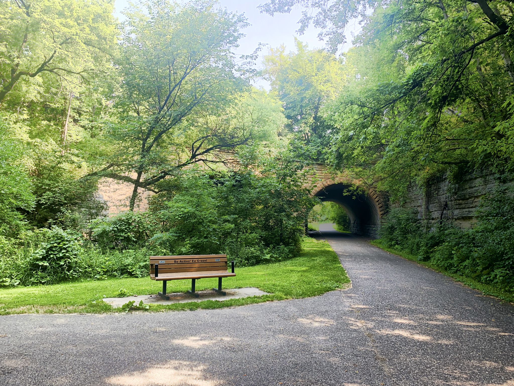 Paved path leading to a stone tunnel, with a bench under dappled sunlight. - Bruce Vento Regional Trail