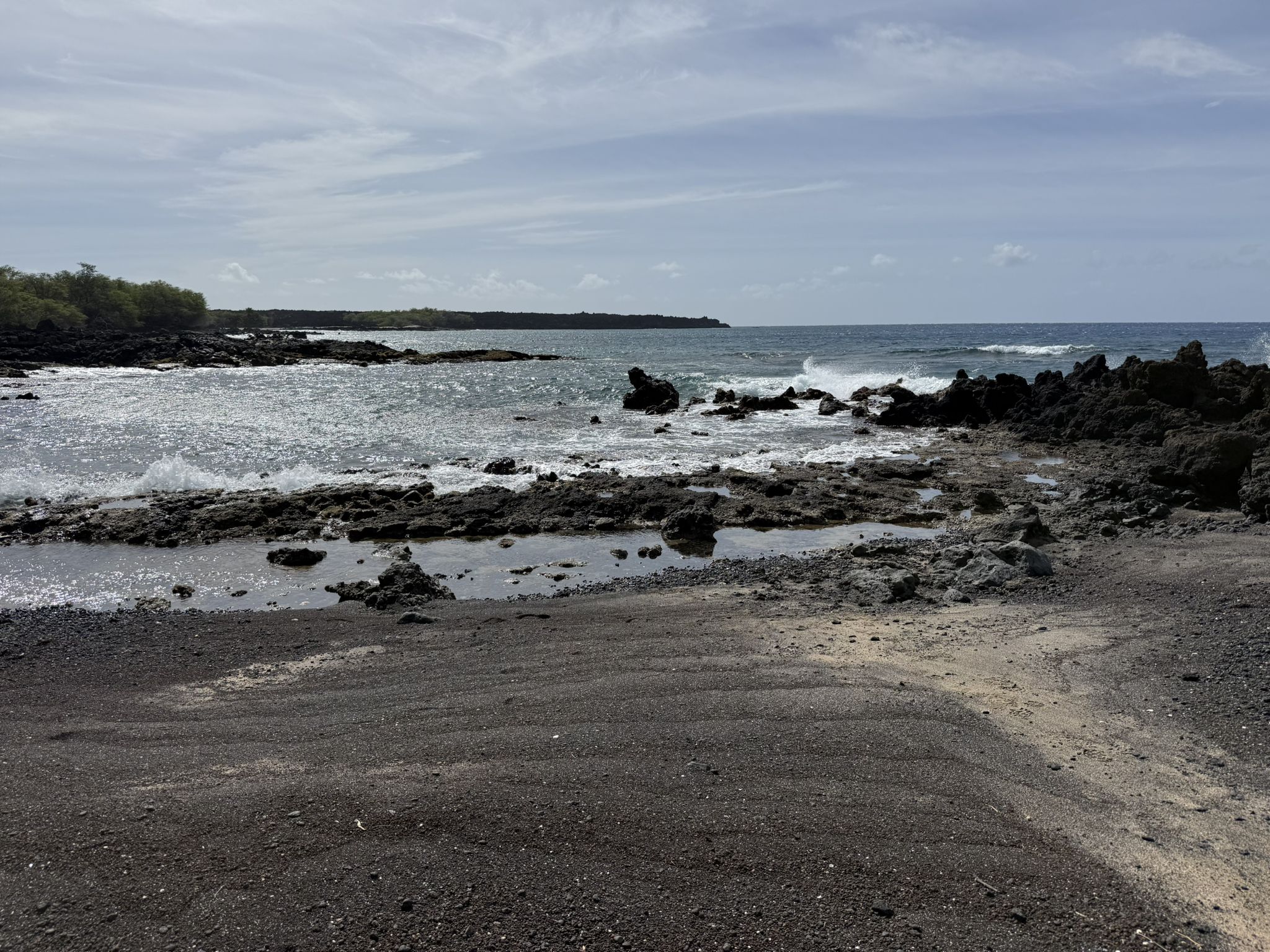 Rocky beach with dark sand, ocean waves, and a cloudy sky. - Hoapili Trail