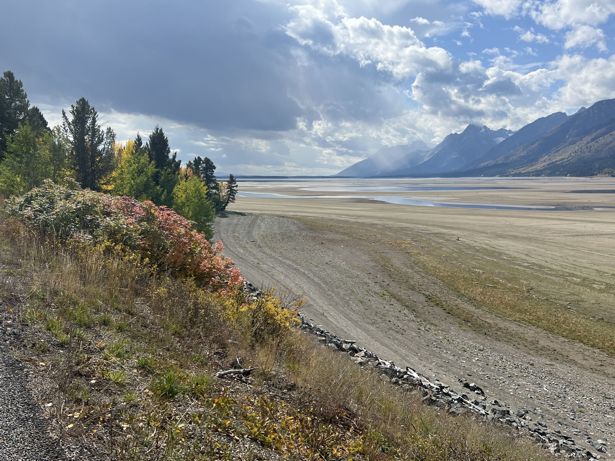 Wide, dry riverbed with autumn trees and distant mountains under cloudy sky. - Grand Teton National Park Road Trip