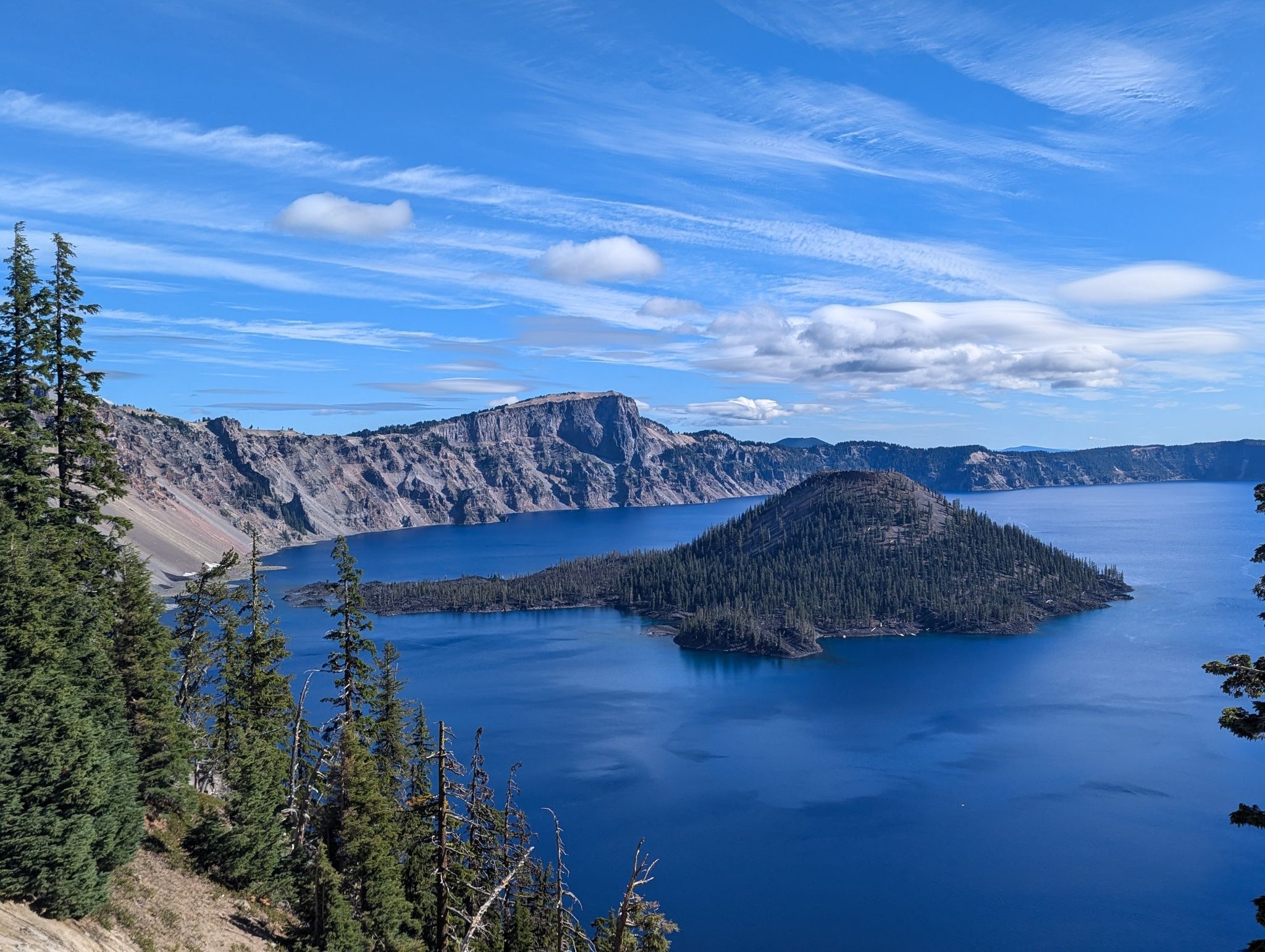 Deep blue lake with a forested island, surrounded by rugged cliffs under a cloudy sky. - Rim Trail and Discovery Point Trail