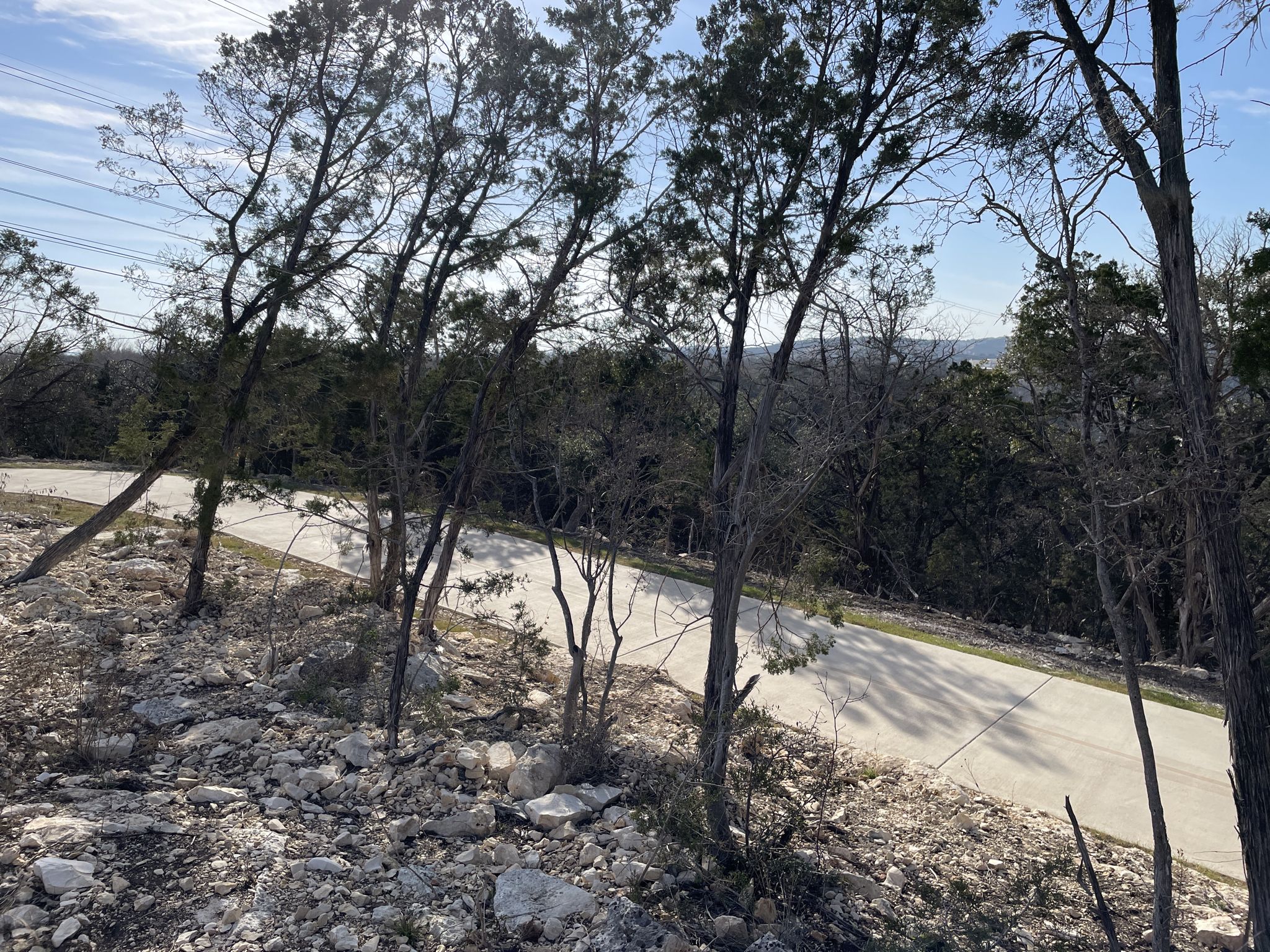 A paved path winds through a rocky, wooded hillside under bright sunlight. - Salado Creek Trail – Morningstar Boardwalk loop from Six Flags Fiesta Texas