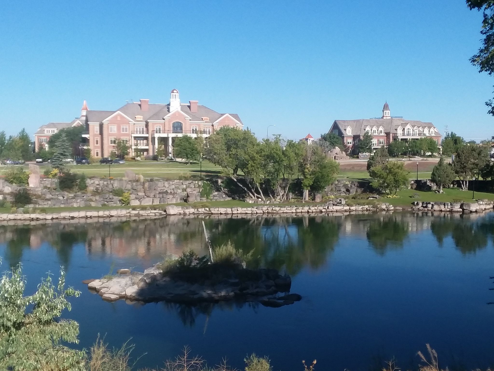 Large buildings reflected in a calm river with a small rocky island. - Friendship Garden – Idaho Falls Greenbelt loop from Hero Arena