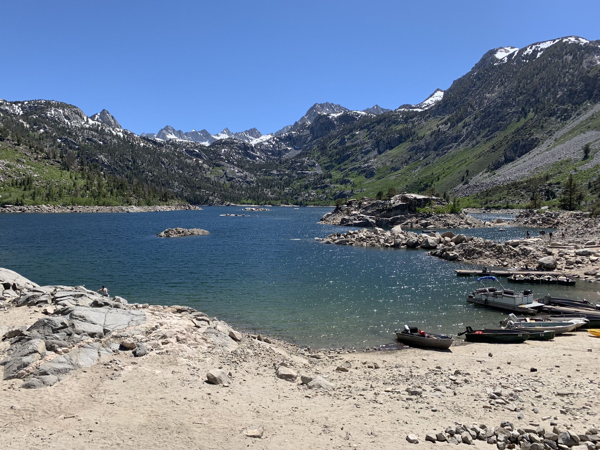 Sandy shore of a large lake with several small boats and snow-dusted mountains. - Sabrina Lake loop from Aspendell