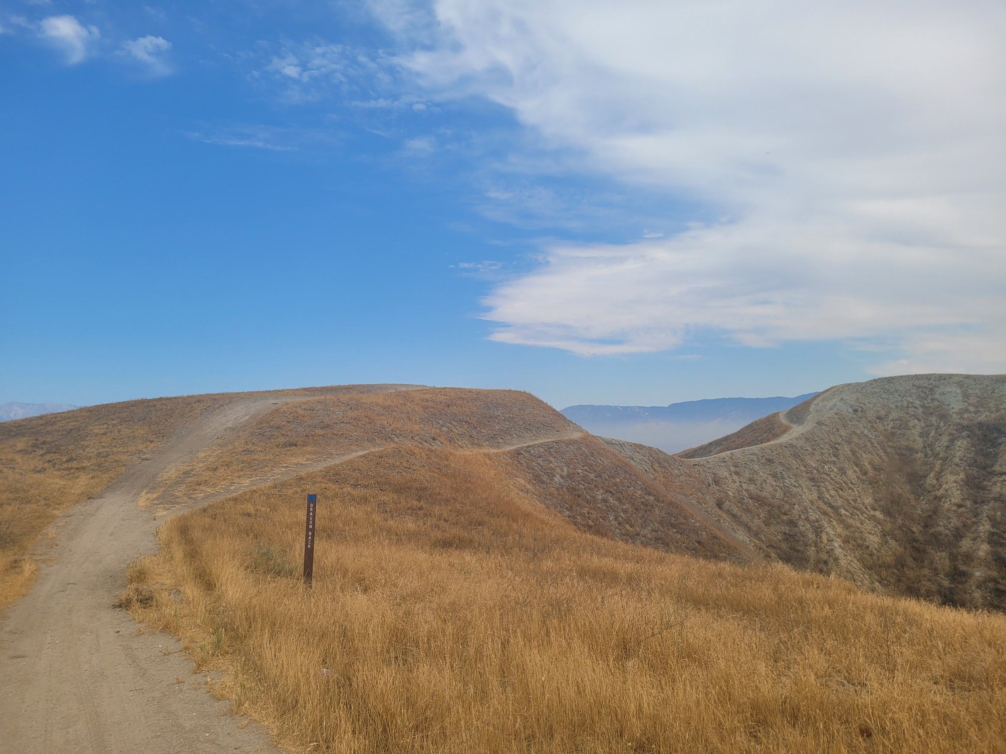 A dirt trail winds through dry, golden hills under a bright, cloudy sky. - Hidden Bear Trail