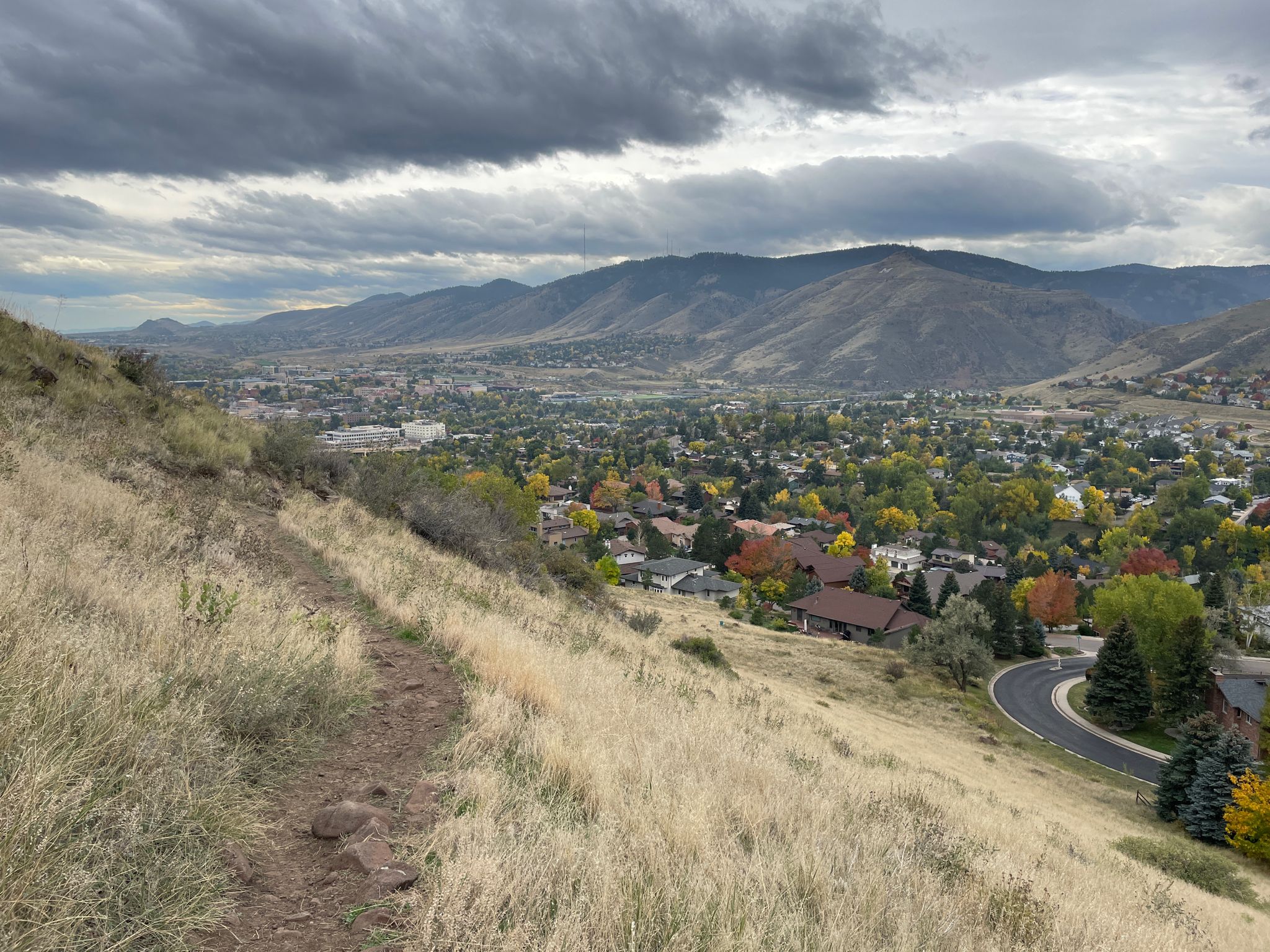 Dirt trail on a grassy hillside overlooking a town and mountains under cloudy sky. - Tilting Mesa Trail – North Table Mountain loop from Golden