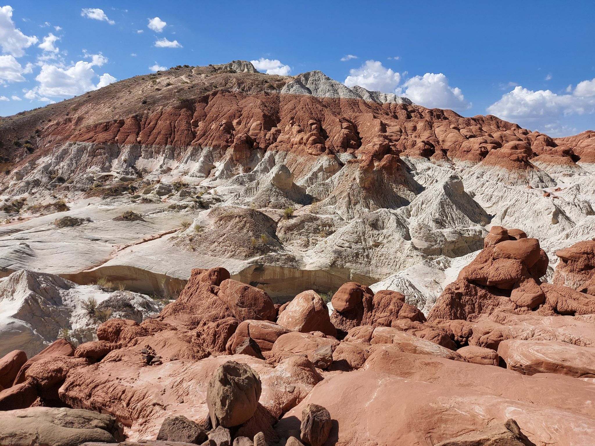 Layered red and white desert hills with scattered rock formations under a partly cloudy sky. - Toadstool Hoodoos Trail