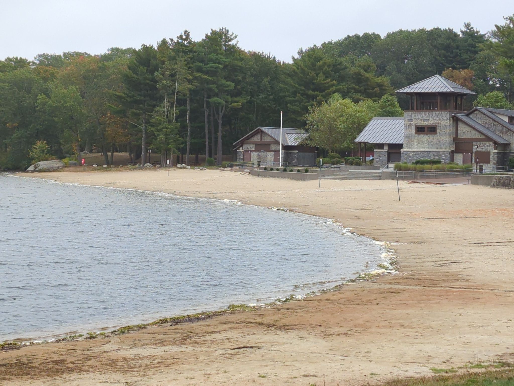 Empty sandy beach curving along a lake, with buildings and trees. - Les Pawson Trail loop