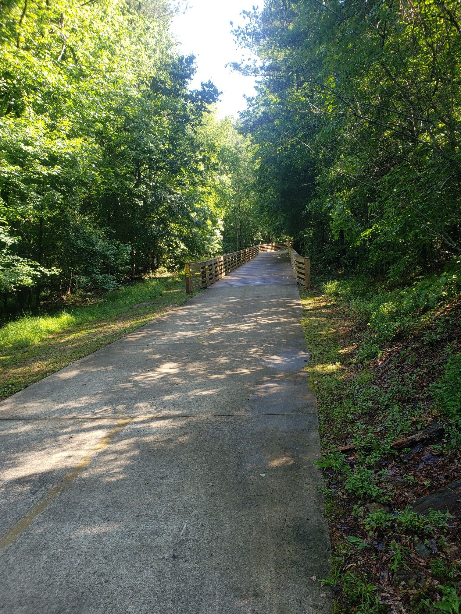 A sunlit paved path leads through green woods towards a distant wooden bridge. - Arabia Mountain PATH Trail