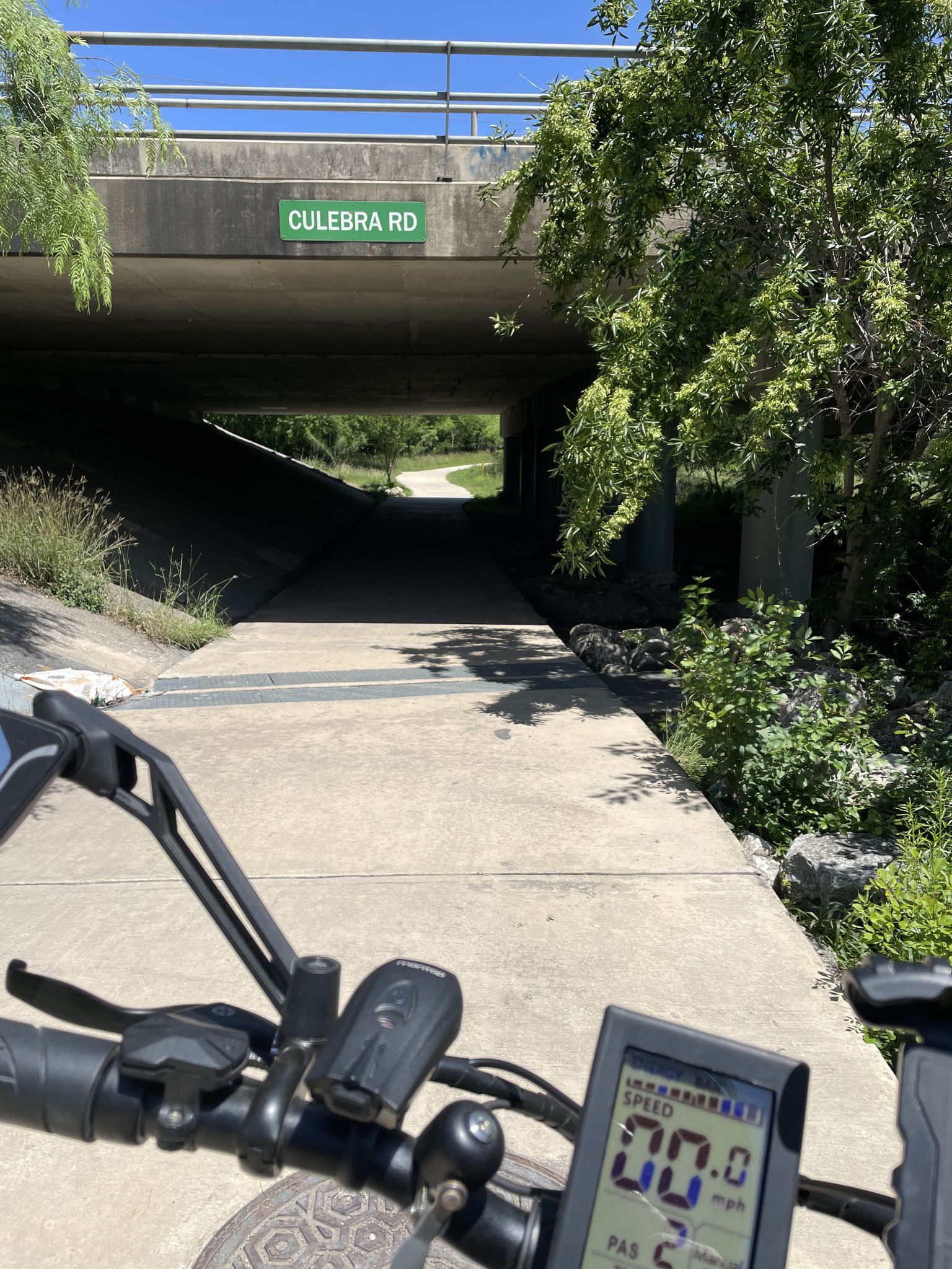 View from a bicycle handlebar of a concrete path leading under a bridge. - Leon Creek Greenway Trail