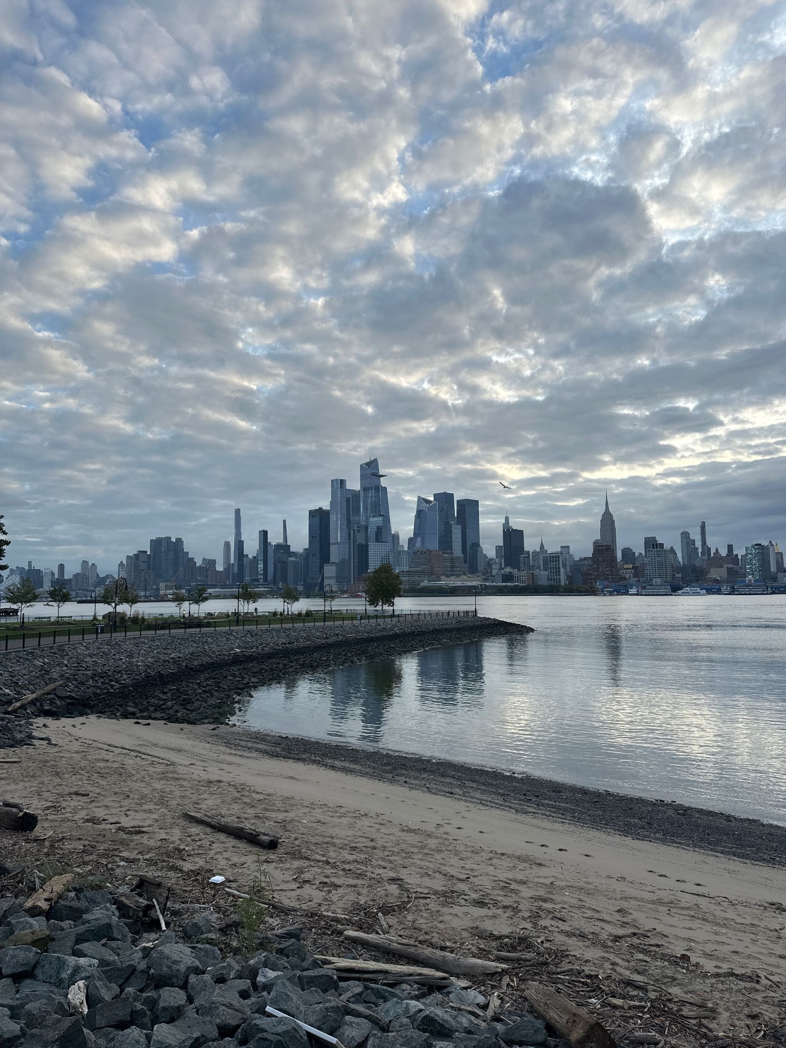 City skyline reflected in calm water, with a sandy and rocky shore foreground. - Hudson River Waterfront Walkway and 9-11 Trail