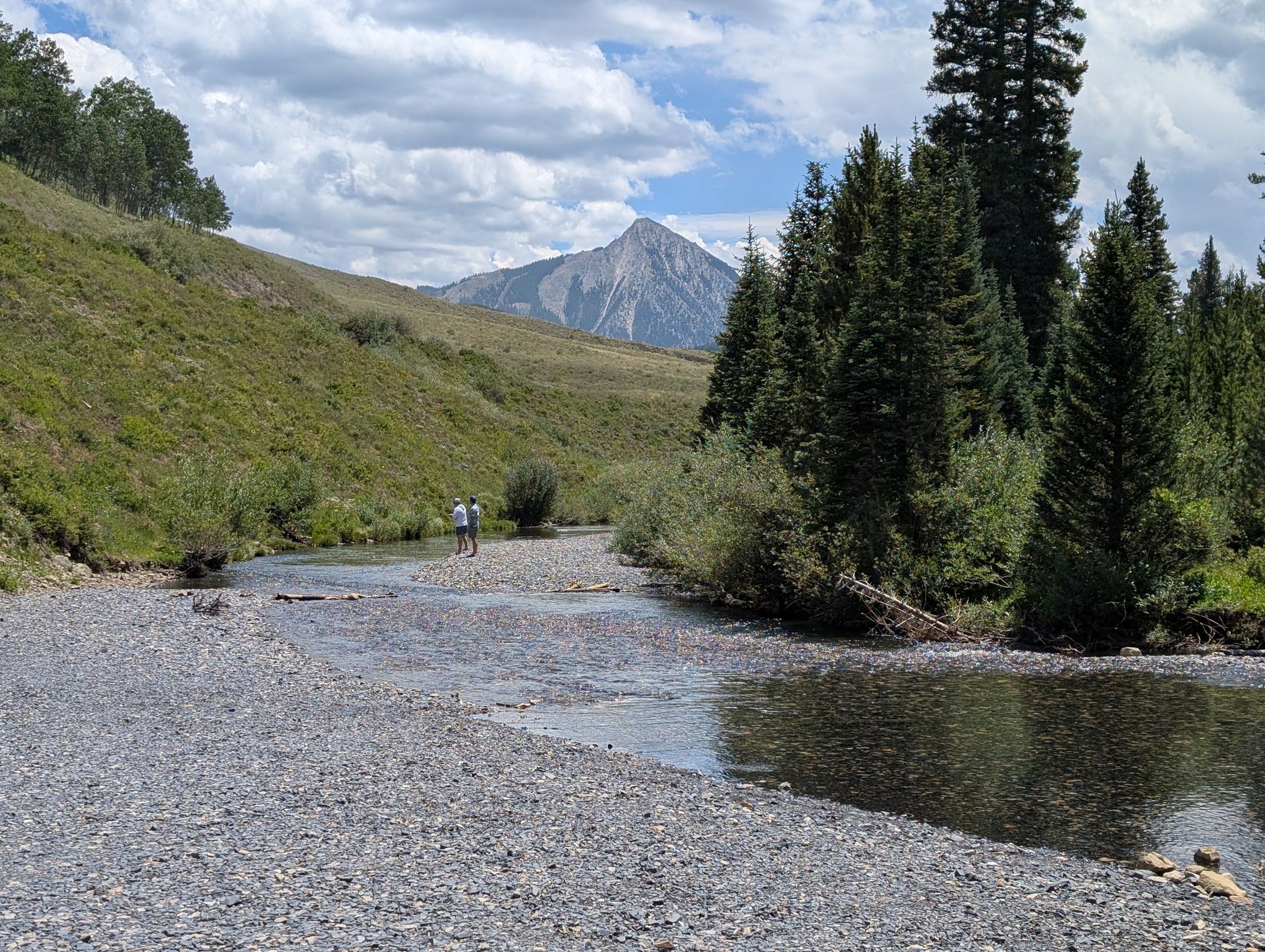 Two people by a rocky river with grassy hills, evergreen trees, and a mountain. - Oh-Be-Joyful and Daisy Pass Loop