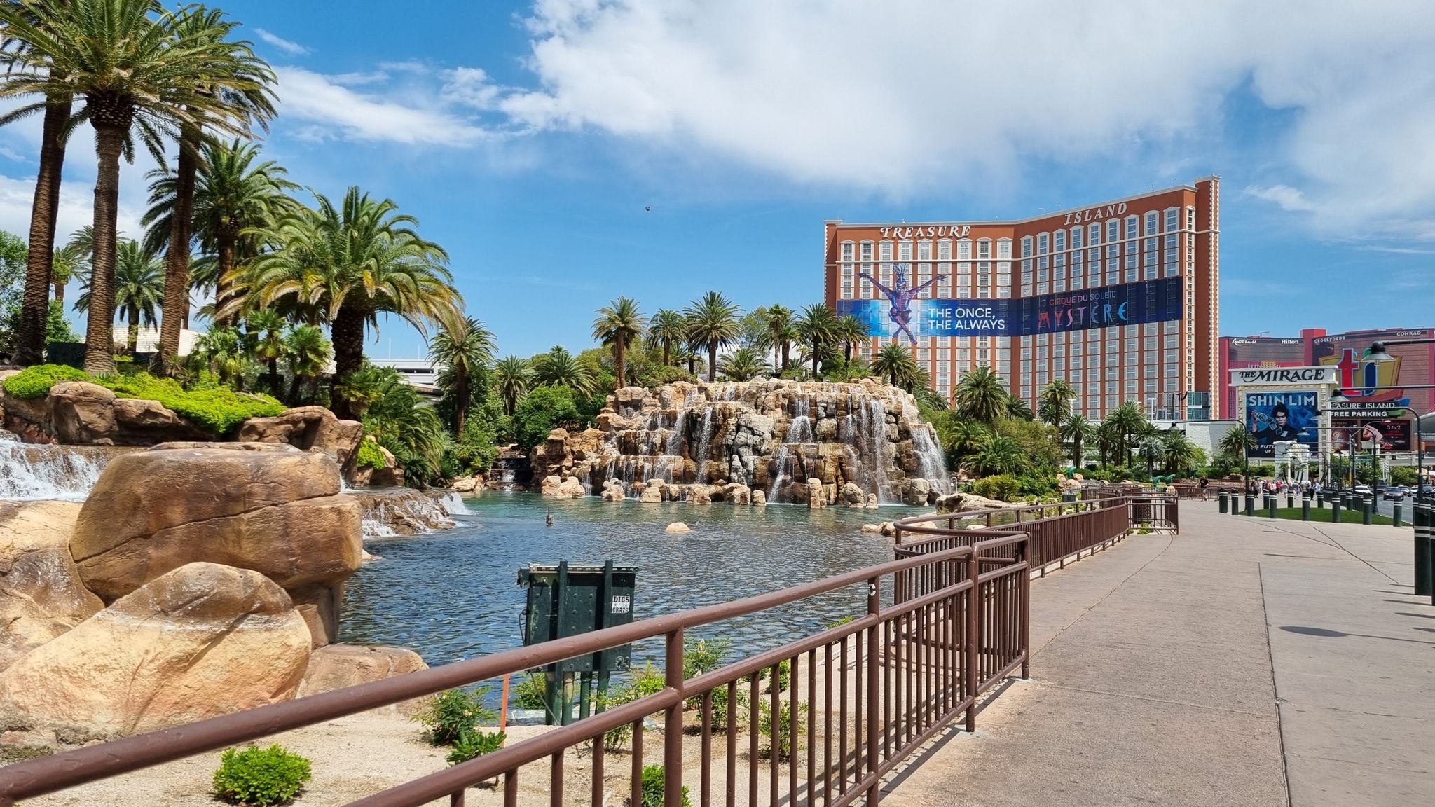 Artificial waterfall and pond with palm trees and a large building. - The Strip