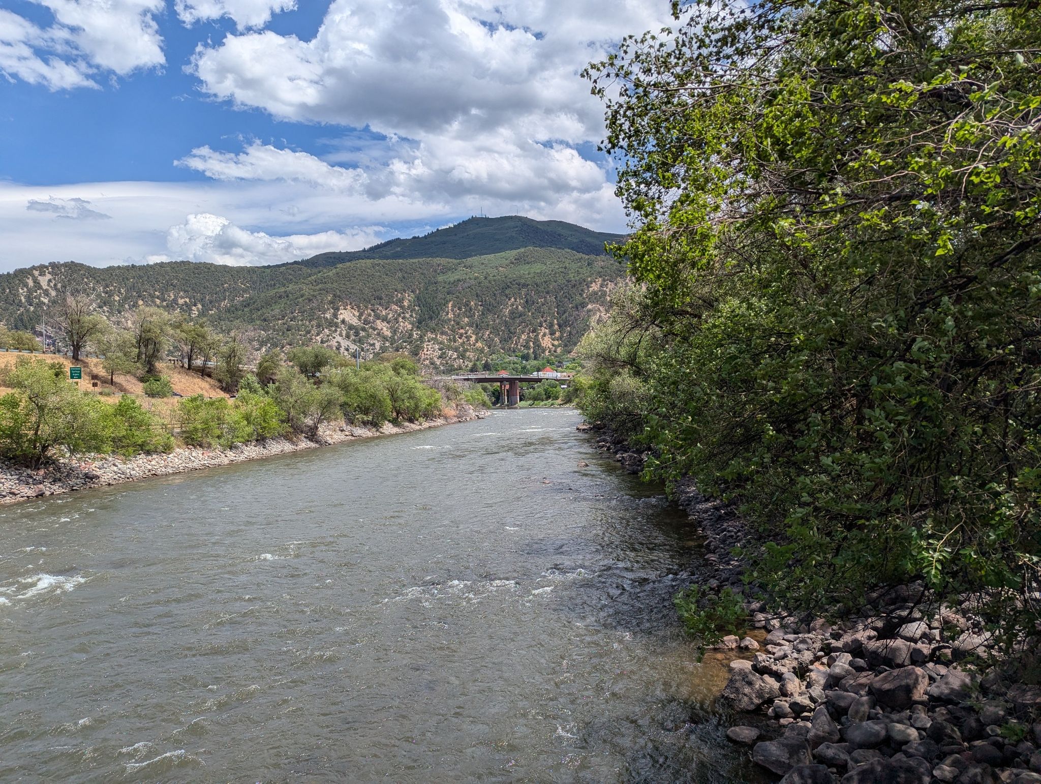 A wide river flows past green trees and distant mountains under a cloudy sky. - View of Roaring Fork River – Two Rivers Park loop from Sopris Elementary School