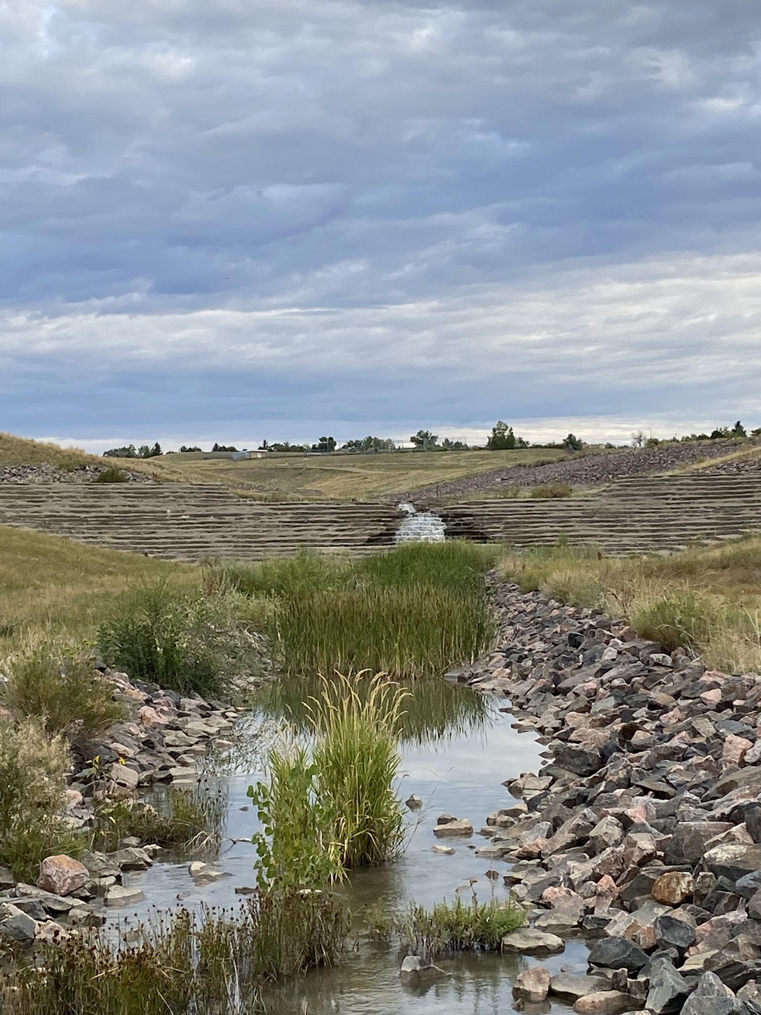 A small stream flows through a rocky, grassy channel under an overcast sky. - Standley Lake Viewing Area – Standley Lake loop from Two Ponds National Wildlife Refuge