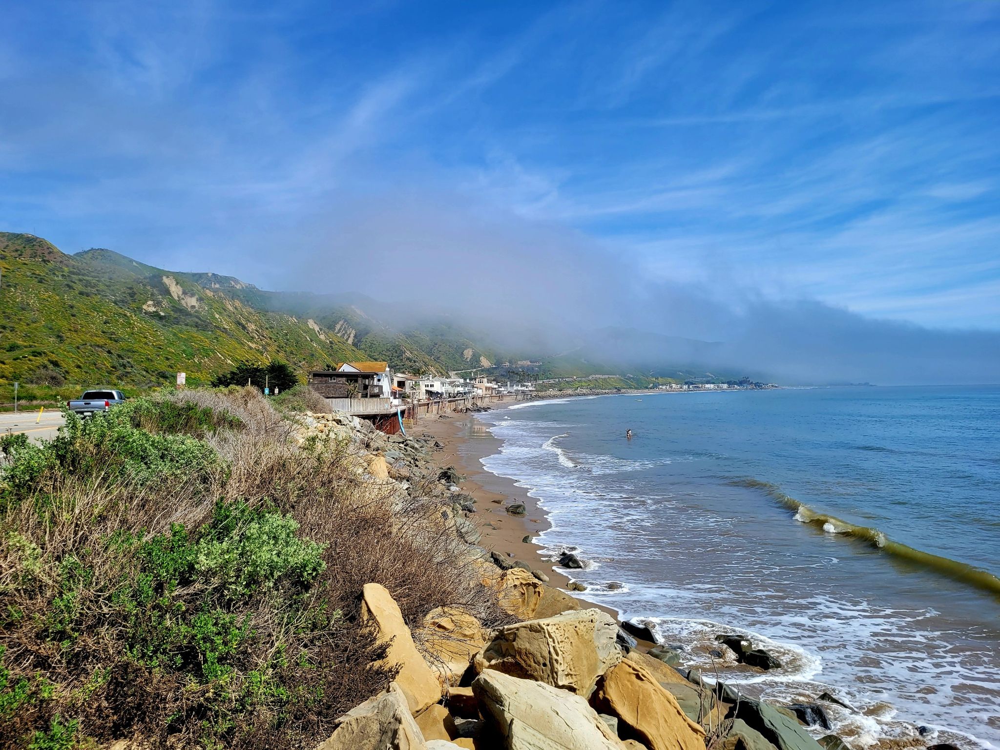 Coastal view with green hills, houses, ocean, and rocks under a bright blue sky. - Omer Rains Coastal Bikeway and Ventura Loop