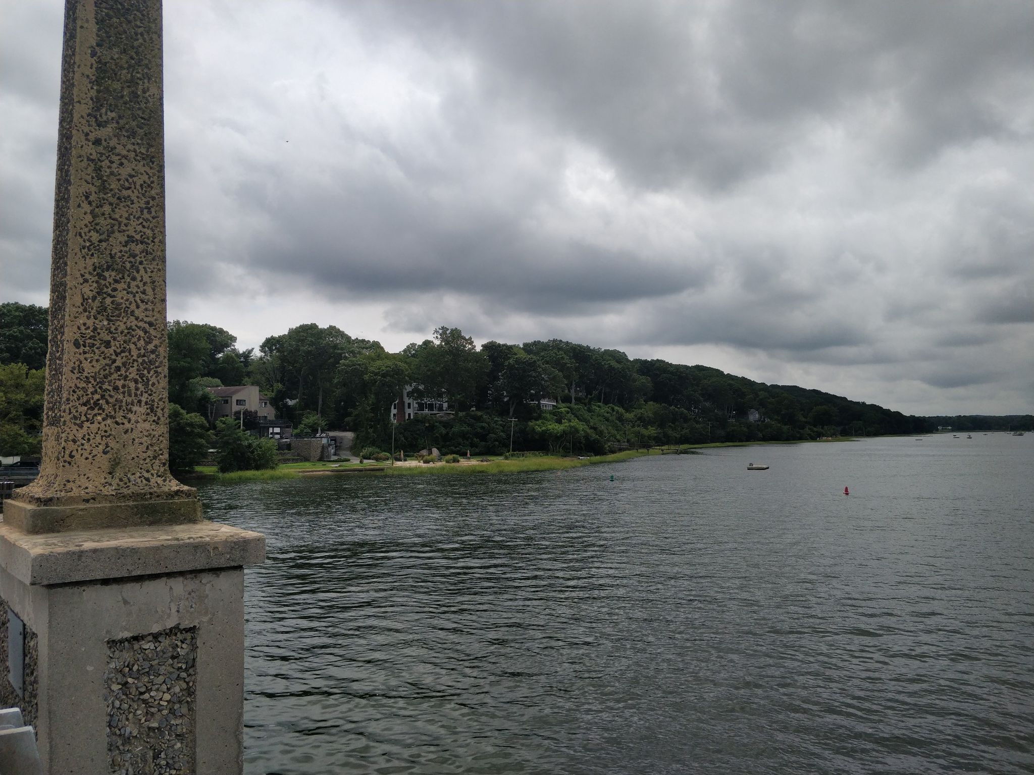 Overcast view of a bay with a forested shore, houses, and a stone pillar. - West Shore Road