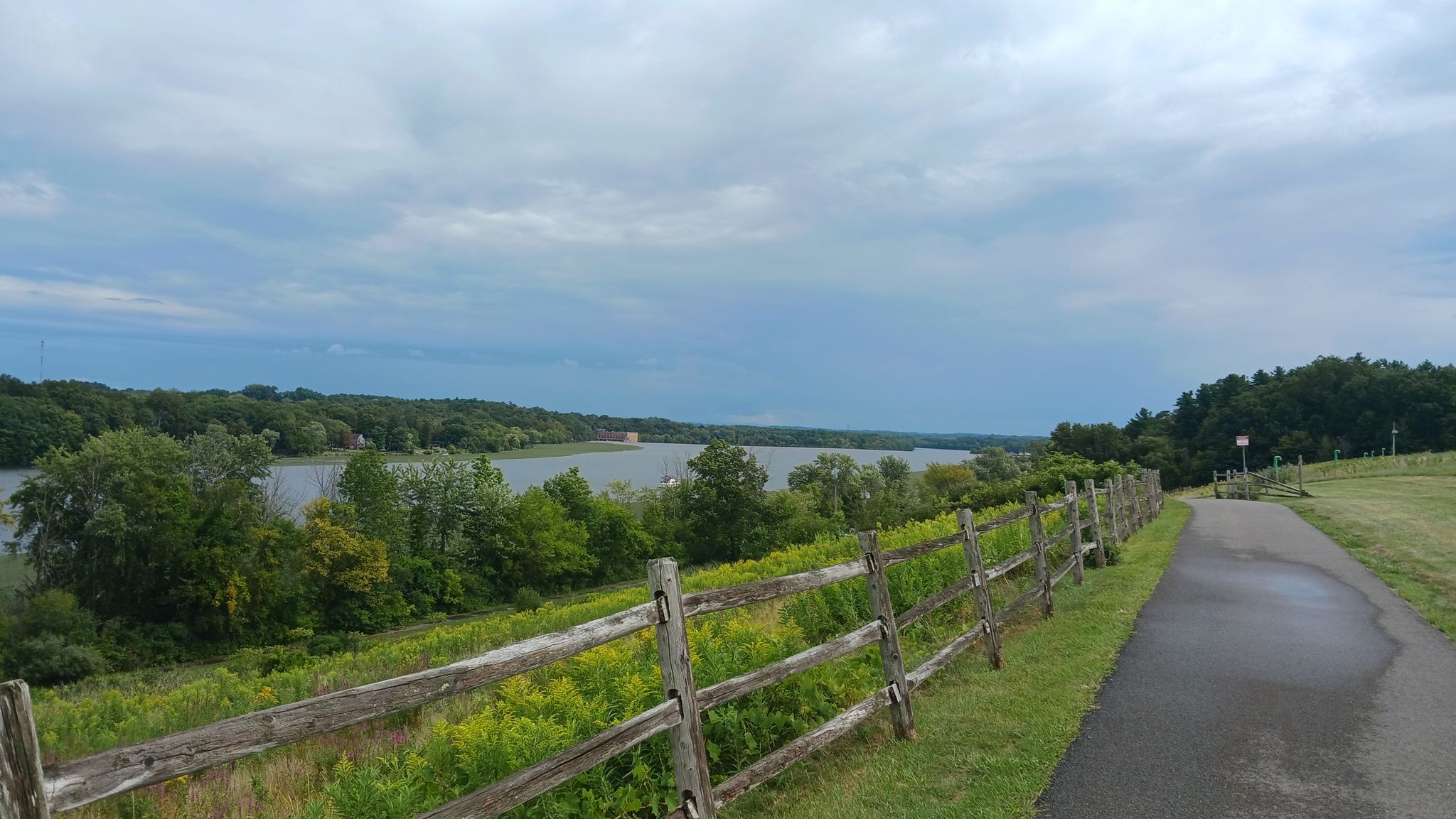 Paved path and wooden fence overlooking a large river under cloudy sky. - View of the Mohawk River – Mohawk-Hudson Bike & Hike Trail loop from Mohawk River State Park
