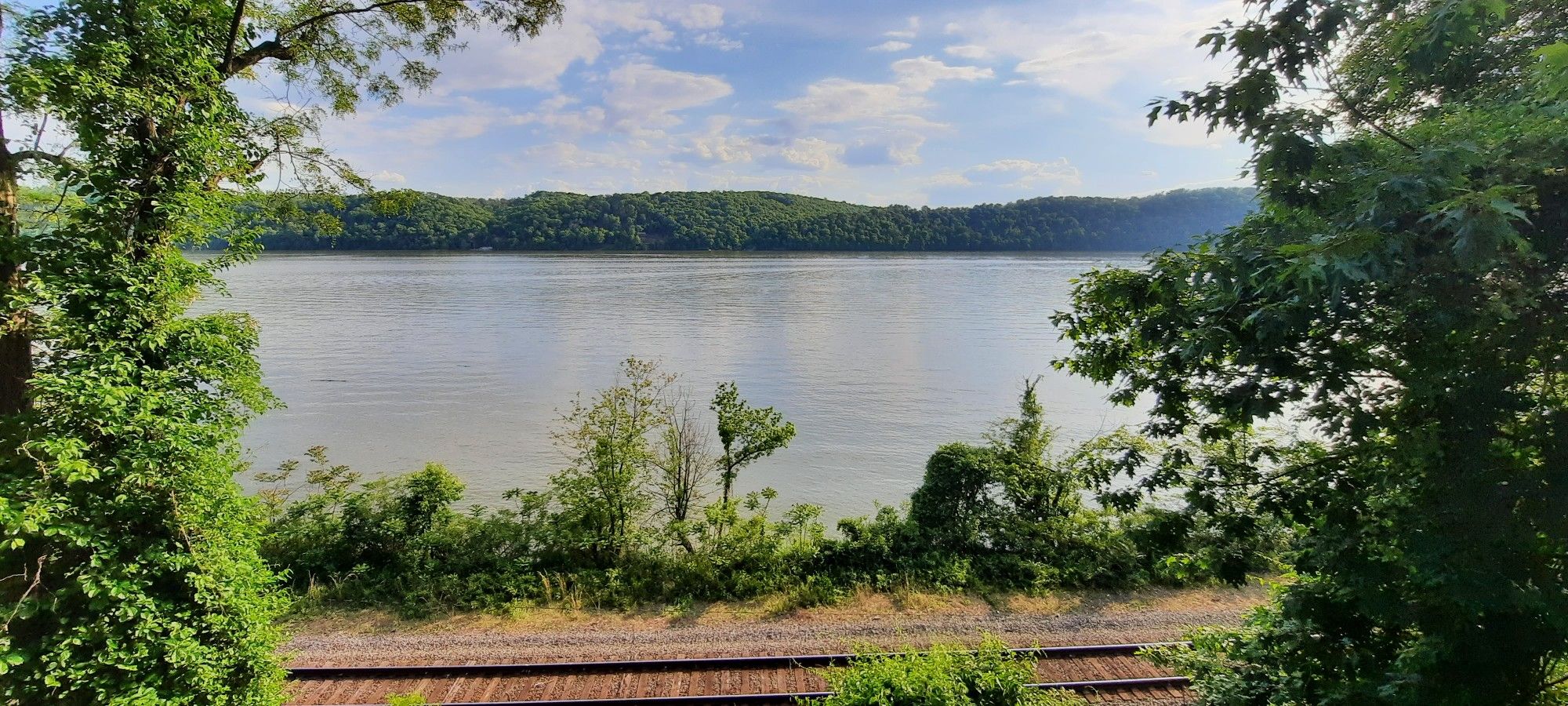 Wide river with forested hills under a bright sky, railroad tracks in foreground. - Enola Low Grade Trail