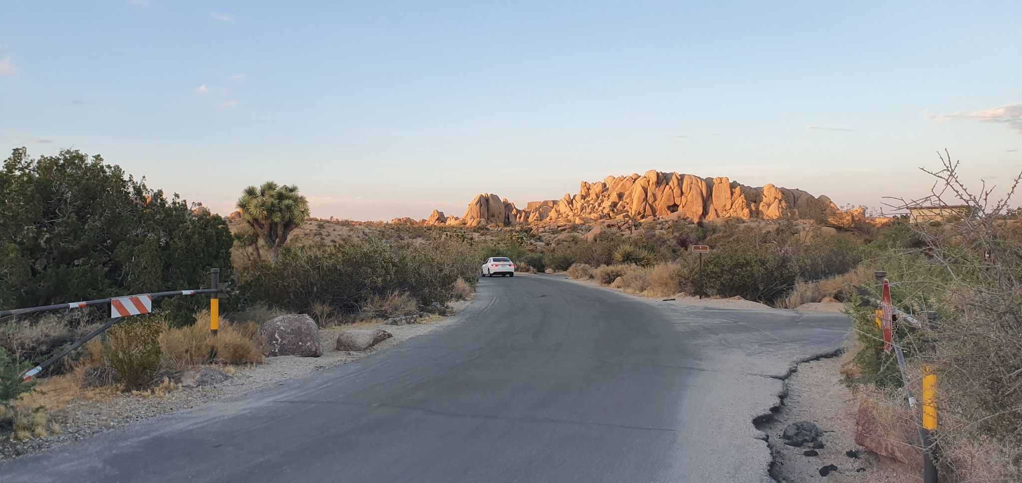 Paved road through desert with a distant white car and rock formations at sunset. - Chimney Rock – Jumbo Rocks Campground loop from Jumbo Rocks