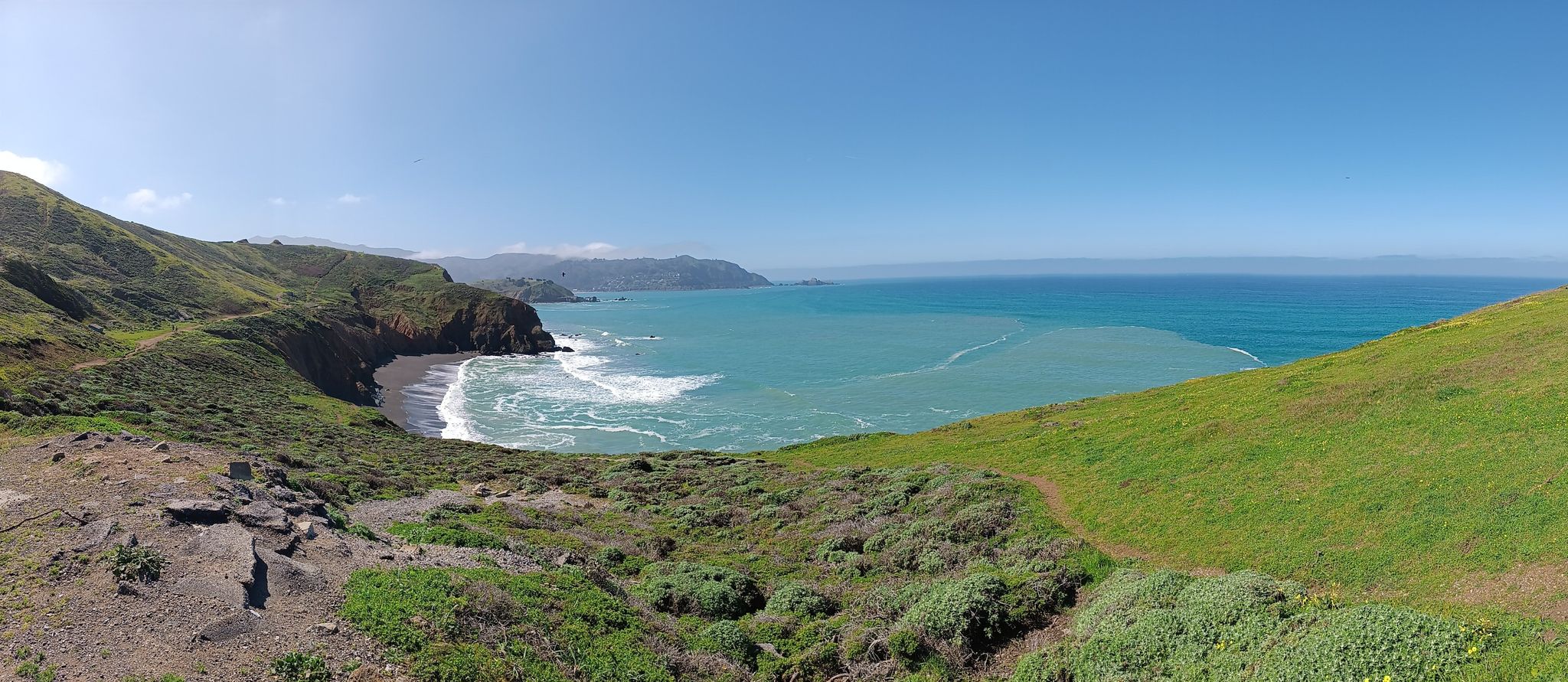 A panoramic view of green coastal hills overlooking a turquoise ocean cove. - Mori Point Loop Trail