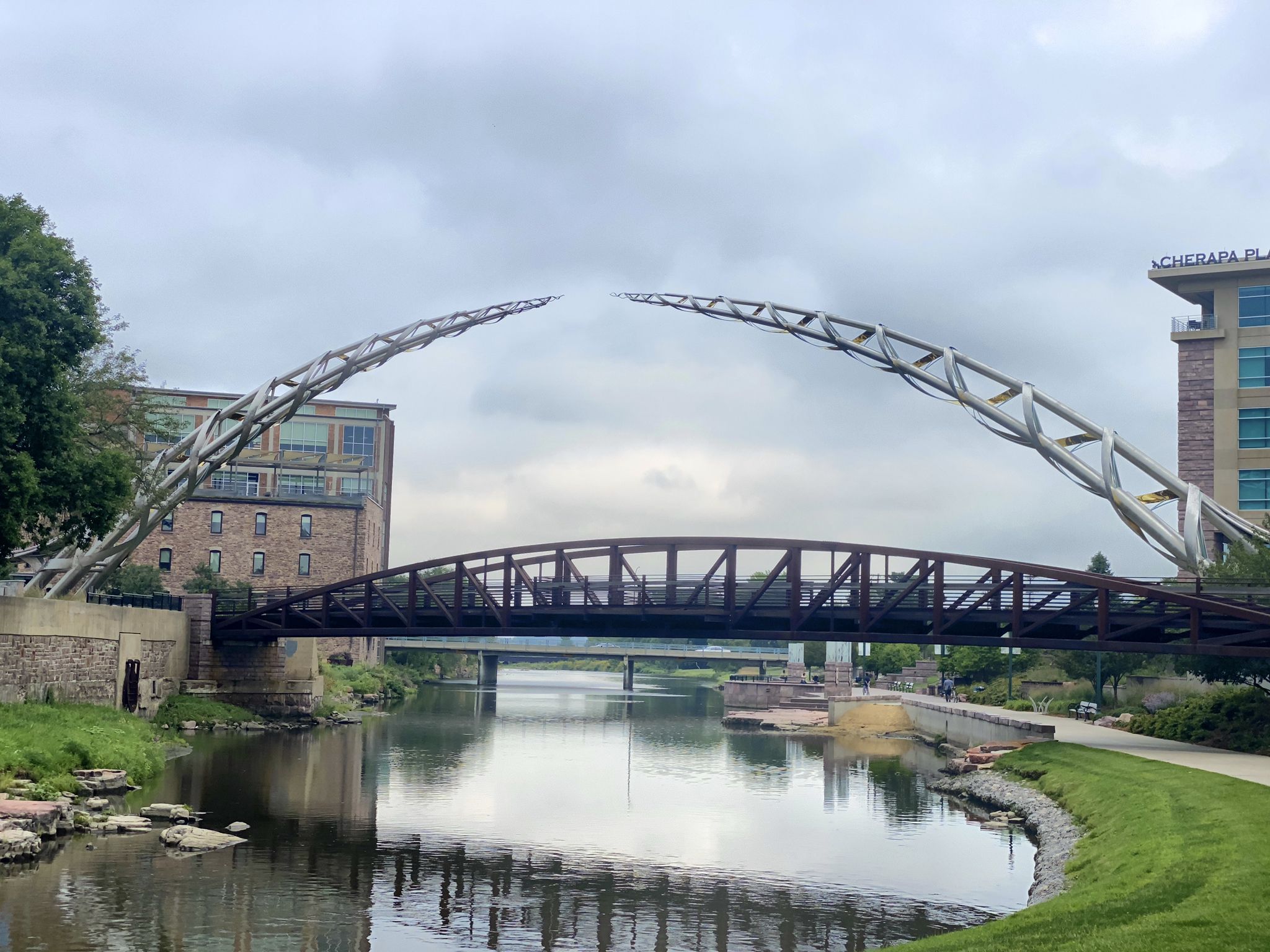 Two modern arched bridges span a river with urban buildings under overcast sky. - Sioux Falls Bike Trail loop from Lincoln High School