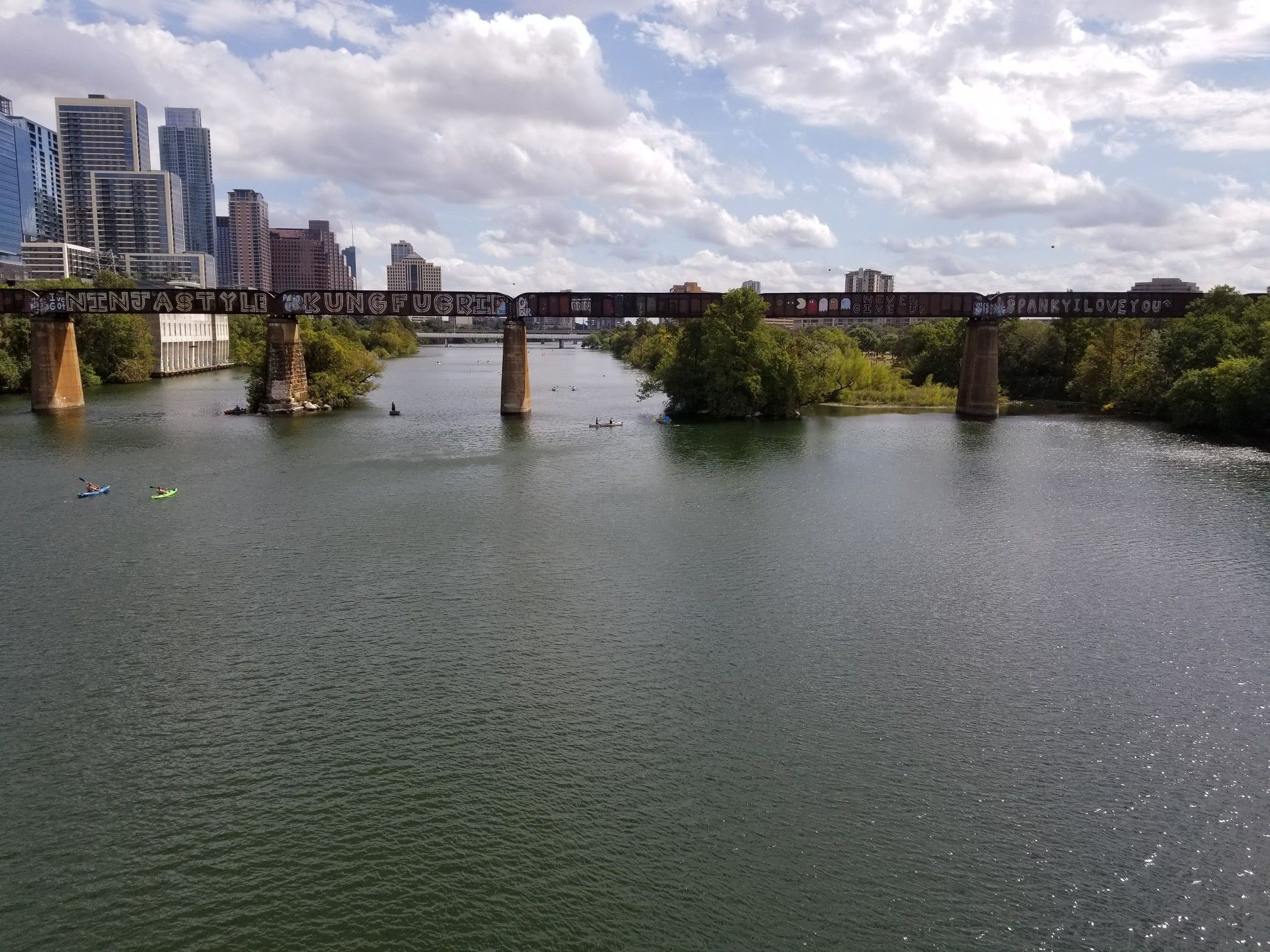 Cloudy day over a wide river, train bridge, distant city, and boats. - Barton Creek Pedestrian Bridge – Lamar Boulevard Bridge loop from Travis