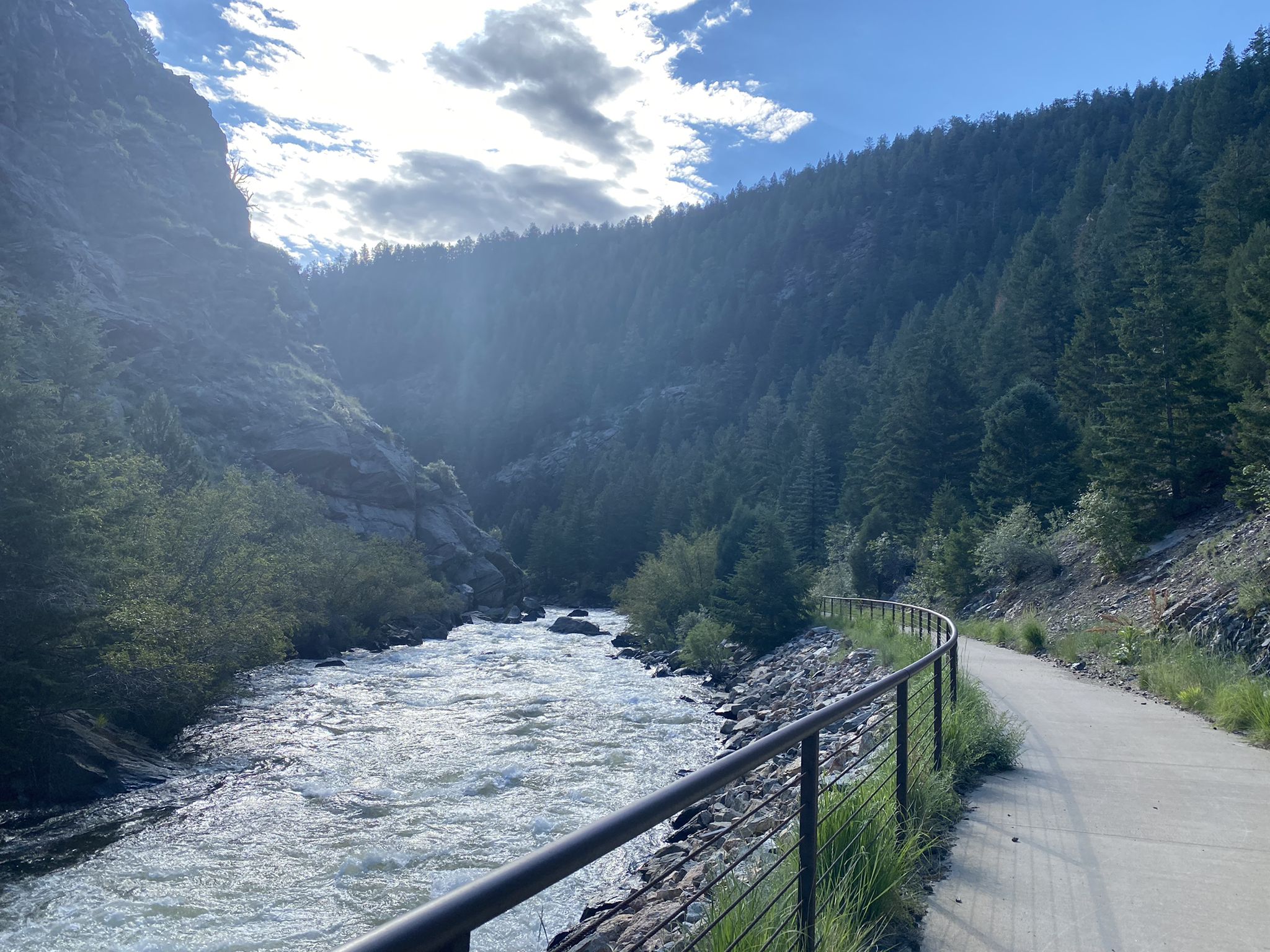 Paved path alongside a river flowing through a sunny, forested mountain valley. - Peaks to Plains Trail
