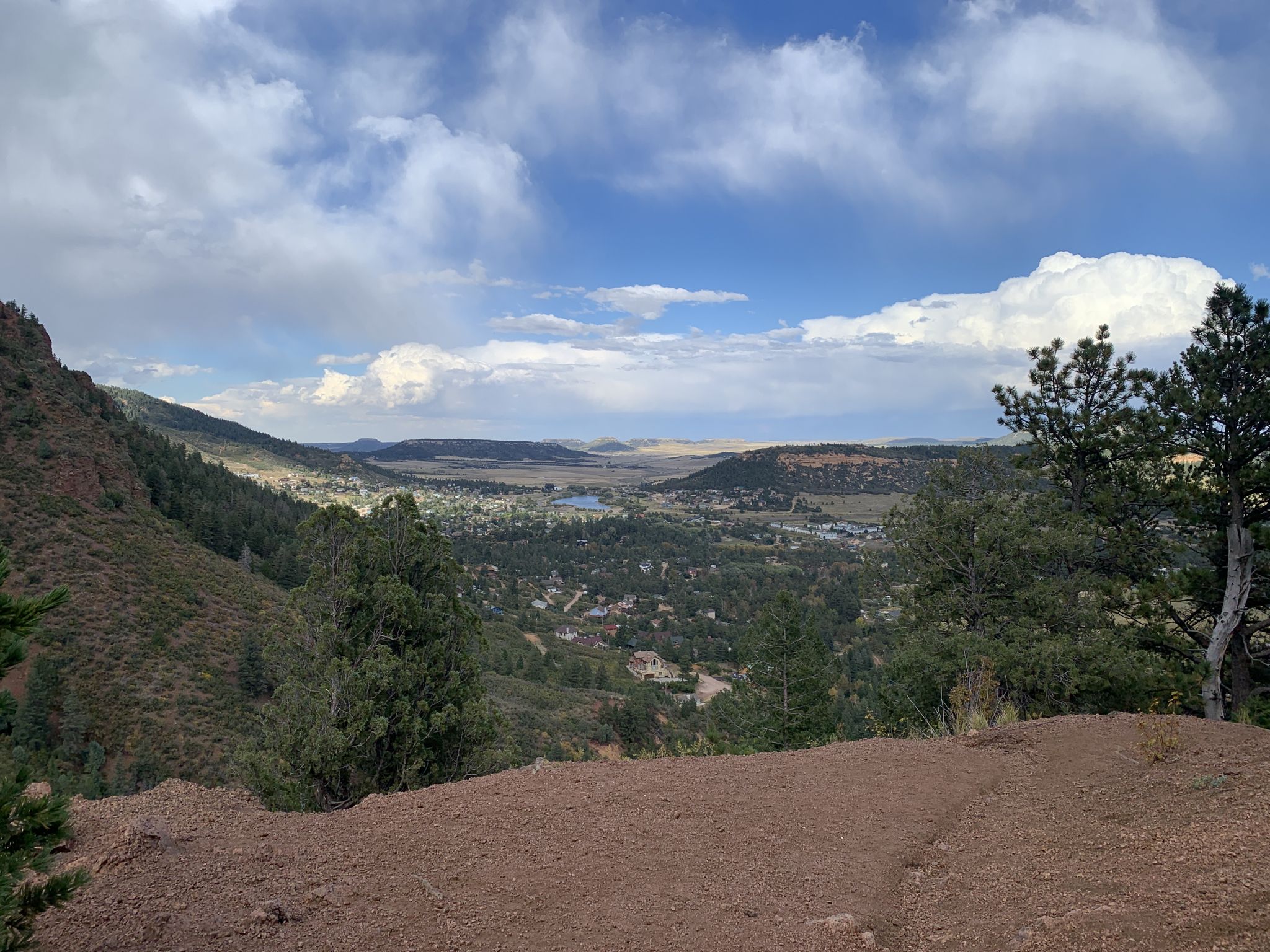 A panoramic view of a valley with a town, lakes, and distant mountains under a cloudy sky. - Raspberry Mountain Loop
