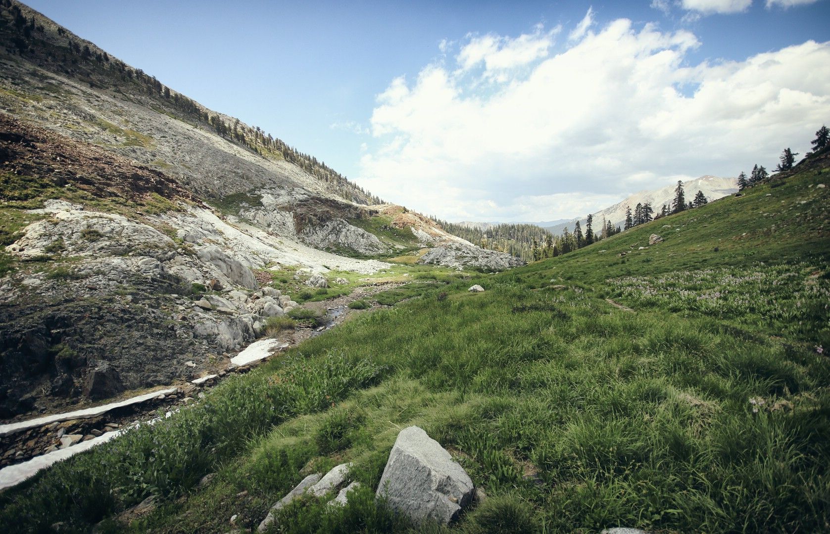 A mountain valley with rocky slopes, green grass, and patches of snow under a cloudy sky. - Mosquito Lakes Trail