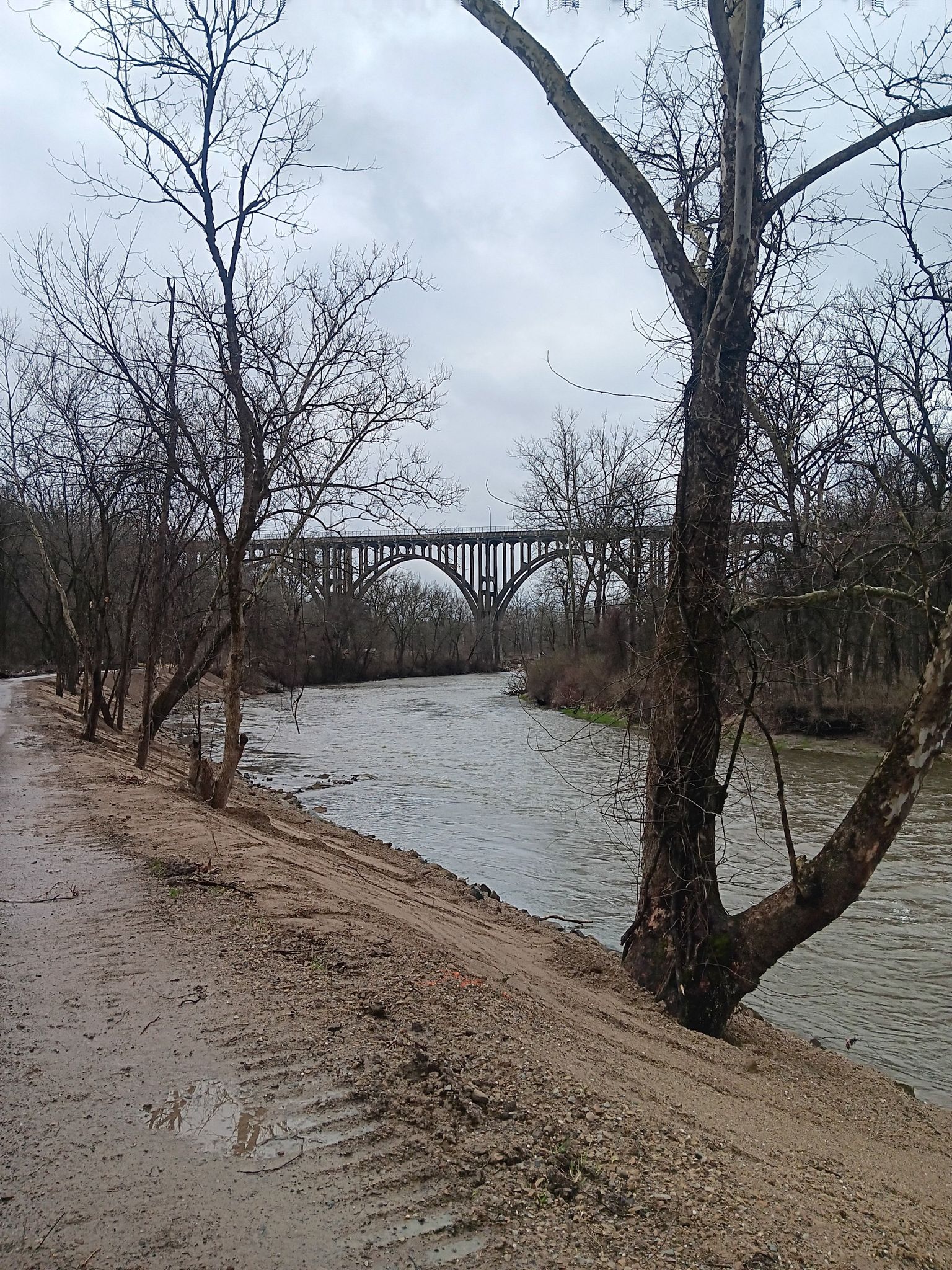 Muddy path next to a wide river, with a large arched bridge in the distance. - Quiet all purpose trail – Brecksville-Northfield Bridge loop from Big Creek & Memphis Railway