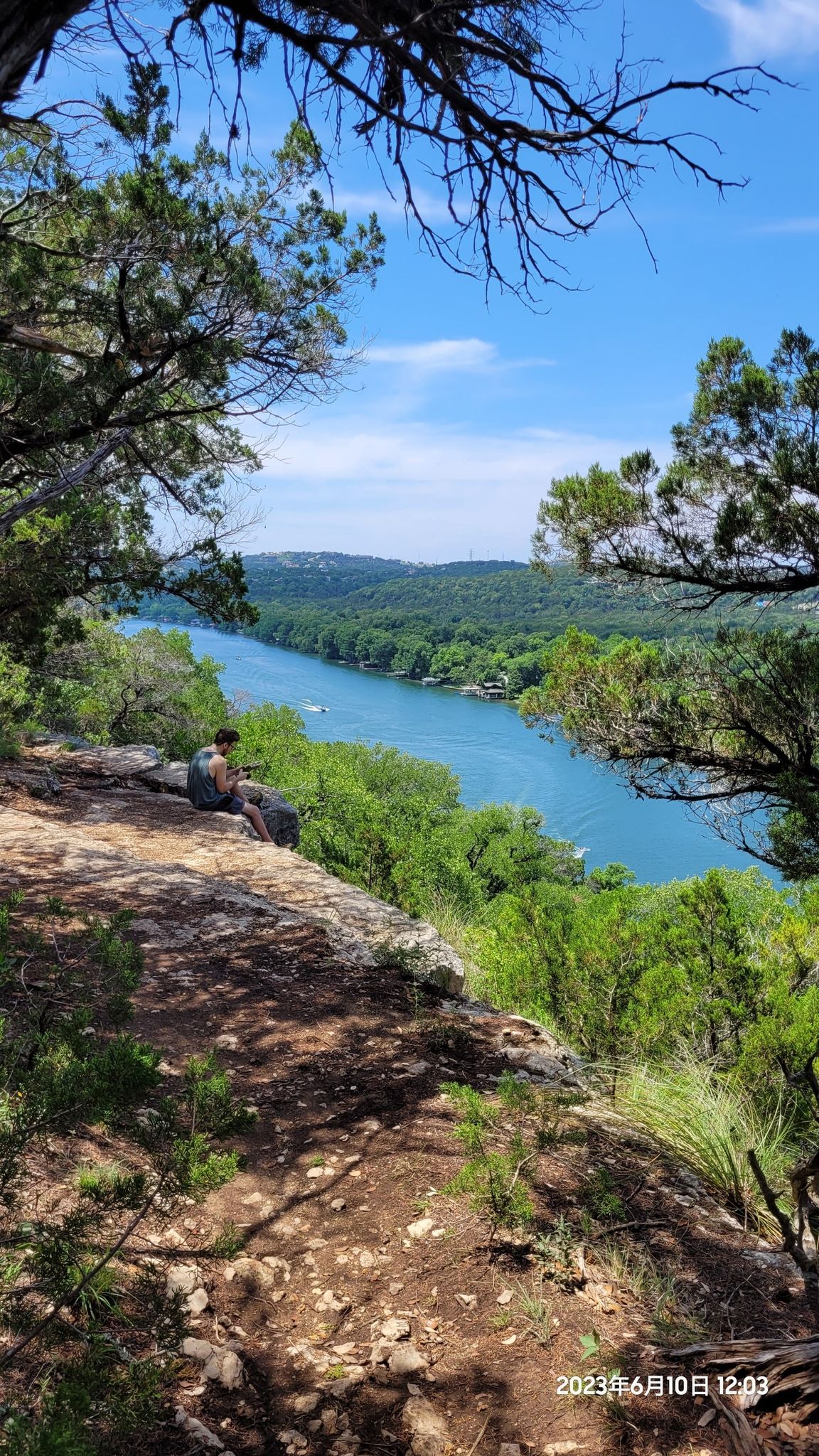 Sunny view of a person sitting on a cliff overlooking a blue river. - Emma Long Cliff Loop