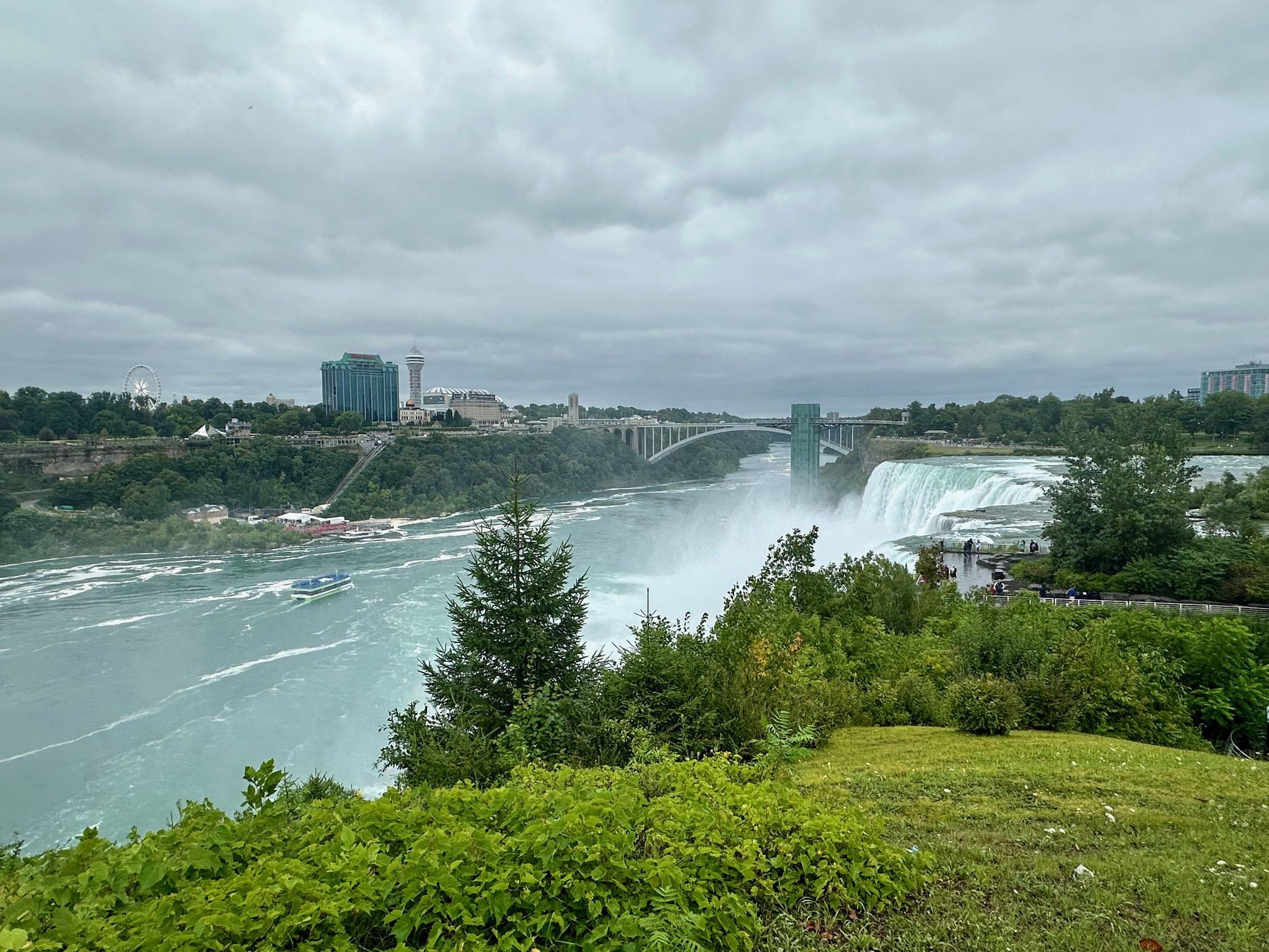 Waterfall, river, and bridge with distant city under a cloudy sky. - Niagara Falls State Park loop