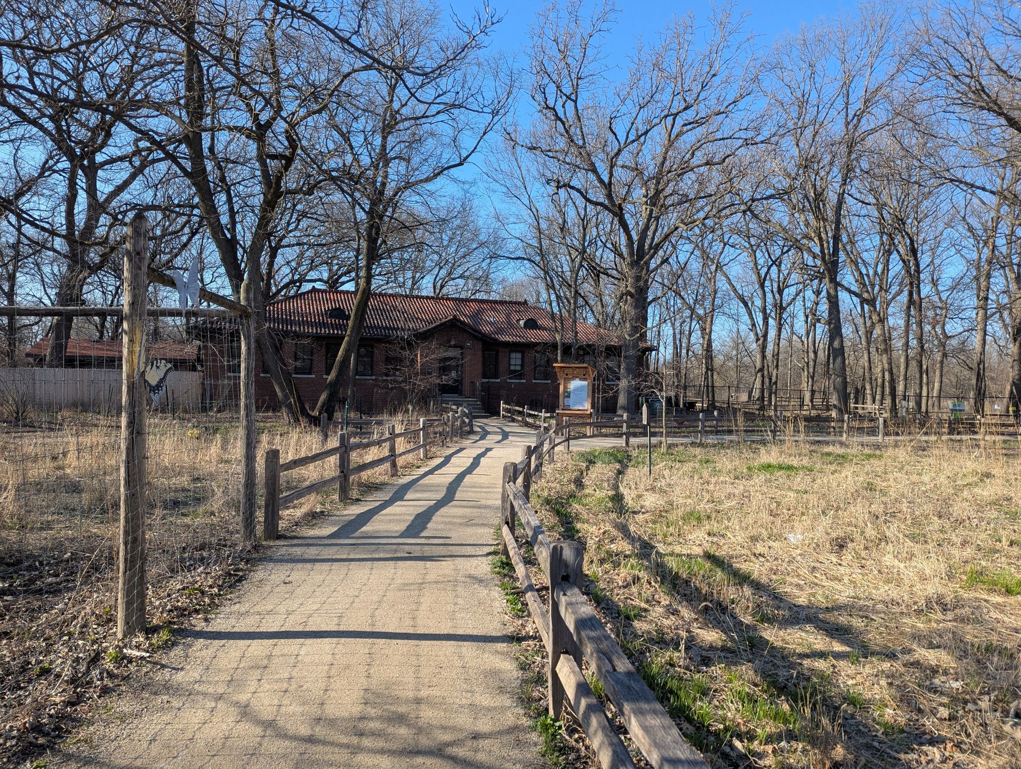 Sunny path leads to a brick building surrounded by bare trees and dry grass. - North Park Village Nature Center Trail